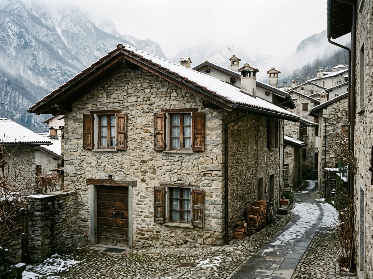 Casa d'epoca lombarda in pietra con infissi datati e neve leggera sul tetto, in un borgo pedemontano sotto la luce invernale